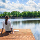 A woman sitting peacefully on a wooden dock, looking out over a calm lake. A notebook is beside her.