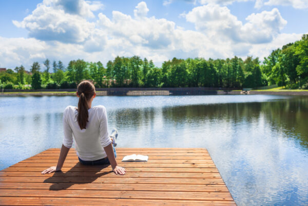 A woman sitting peacefully on a wooden dock, looking out over a calm lake. A notebook is beside her.