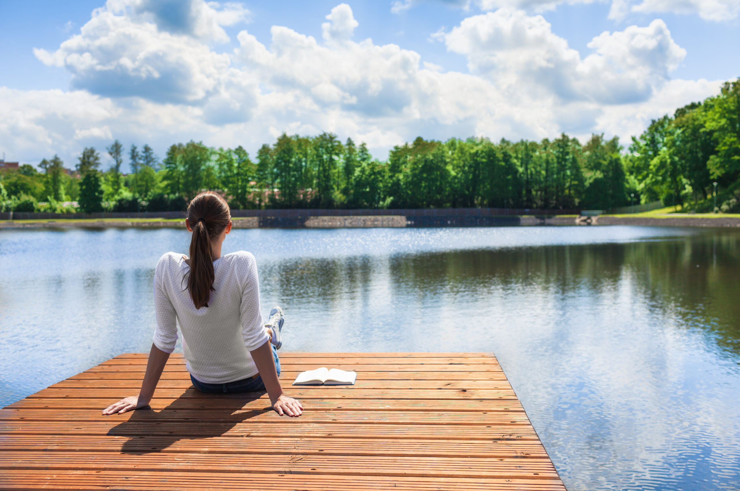 A woman sitting peacefully on a wooden dock, looking out over a calm lake. A notebook is beside her.